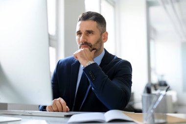 İş planlama konsepti. Düşünceli orta yaşlı işadamı bilgisayarla masada oturuyor, işyerinde çalışıyor ve düşünceli bir ifadeyle monitöre bakıyor.