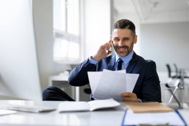 Orta yaşlı işadamı cep telefonuyla konuşuyor ve kağıtları kontrol ediyor, işyerinde oturuyor, finansal raporları okuyan girişimci ve iş ortağıyla telefon görüşmesi yapıyor.