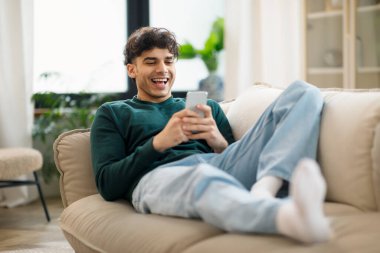 Mutlu Orta Doğulu genç adam cep telefonu kullanıyor ve gülüyor. Evdeki koltukta uzanıyor. Neşeli Adam Akıllı Telefon Oynuyor ve Çevrimiçi Ağ Hafta Sonu Eğleniyor