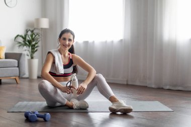 Neşeli, sportif, güzel, esmer, doğulu spor elbiseli, yoga minderinde oturan, omuzunda havlu olan, elinde su şişesi olan, evde egzersiz yapan, uzay fotokopisi çeken bir kadın.