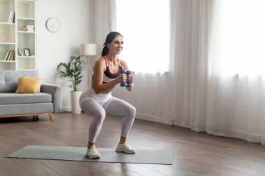 Spor, spor ve sağlıklı yaşam tarzı konsepti. Spor kıyafetleri giymiş, elinde dambıllar tutan, ağırlıklarla evde egzersiz yapan, fotokopi odasına bakan atletik gülümseyen esmer bir kadın.