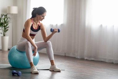 Güçlü, sportif, genç, güzel, sağlıklı bir vücudu olan, spor topu üzerinde oturan, ağırlık pompalayan, evdeki dambıllarla egzersiz yapan, boşluğu kopyalayan bir kadın.