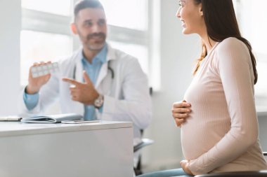 Hamilelik ve ilaç. Erkek uzman hamile kadına hap veriyor, vitamin yazıyor, seçici odaklanıyor. Kadın hasta, hastalığı tedavi etmek için doktordan reçeteli ilaç alıyor.
