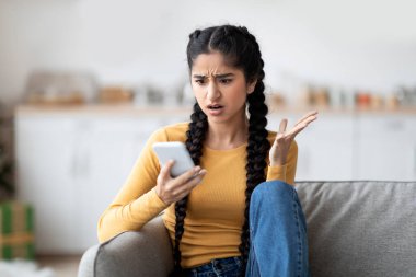 Sinirli genç Hintli kadın evde koltukta oturmuş akıllı telefon ekranına bakıyor. Endişeli doğu hanımı cep telefonundan kötü mesaj okuyor. Haberlere duygusal tepki veriyor.