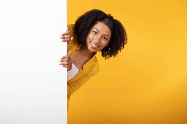有趣的提议 快乐的黑人女士从白板侧面窥视你的文字或设计 兴奋的女人站在黄色背景的广告牌旁边 — 图库照片