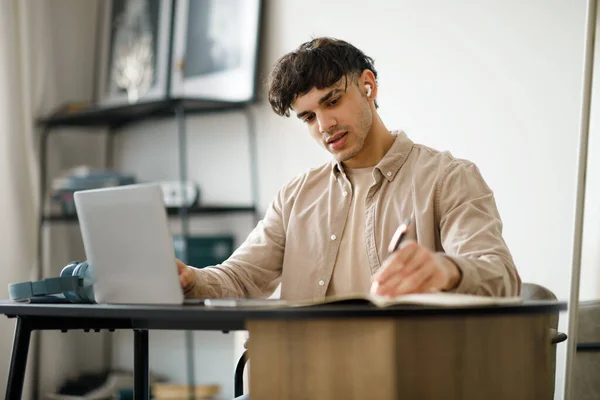 Middle Eastern Guy Using Laptop Websurfing And Taking Notes Sitting At Table At Home. Young Man Learning And Working Online Via Computer Wearing Earbuds At Workplace. Business Study Concept