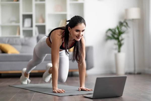 Online fitness classes concept. Beautiful motivated young brunette woman in sportswear exercising on yoga mat in living room, sporty lady have workout at home, using laptop, copy space