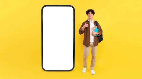 大学生男子拥有巨大的智能手机广告空应用程序屏幕 在黄色工作室背景下批准带Thumbs向上手势的移动提供 — 图库照片