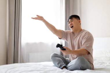 Video oyunu bağımlılığı. Kızgın, orta yaşlı, pijama giyen, elinde kumanda kolu tutan, el kol hareketi yapan, bağırıp çağıran, evde video oyunu oynayan, uzay kopyalayan Asyalı bir adam.