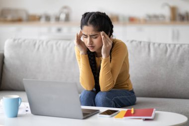 Serbest Stres. Evde dizüstü bilgisayar başında çalışan yorgun Hintli kadın, tapınakları ovuşturan, kaşlarını çatan, oturma odasında otururken şiddetli migren ağrısı çeken genç Doğu 'lu kadın.