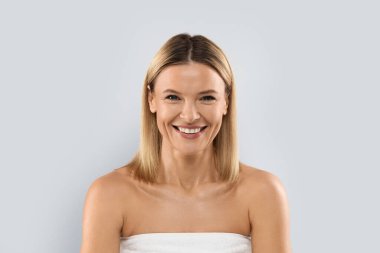 Gülümseyen, orta yaşlı, güzel, sarışın bir kadın. Mükemmel pürüzsüz bir yüzü var. Gri stüdyo arka planında yalnız poz veriyor. Estetik tıp, kozmetoloji konsepti