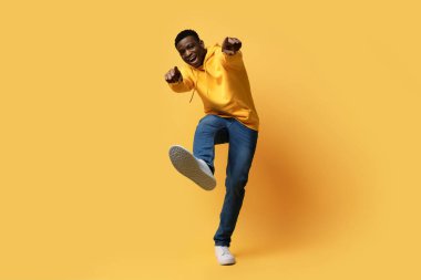 Sarı stüdyo arka planında parmak uçlarını ve ayakkabı ayakkabılarını gösteren şık giysili mutlu, kıvırcık, genç, siyah bir adam, sanki bir şey ya da biri, kopyalama alanı gibi. İnsan jestleri kavramı
