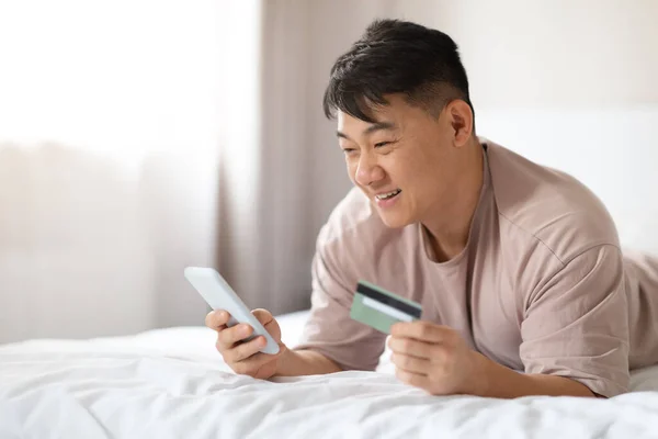 电子商务 虚拟银行业务 快乐而英俊的亚裔中年男子躺在床上 用智能手机和塑料银行卡 在周末呆在家里上网下订单 复制空间 — 图库照片