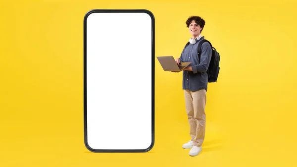 教育应用 快乐学生使用笔记本电脑投递巨大的手机空白屏幕 广告移动应用程序和提供站在黄色背景 Mockup Panorama — 图库照片