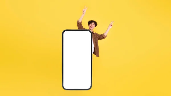 陽気若い男は 大きなガジェットの後ろに立って スタジオで手を上げる 黄色の背景に空の画面にオーバーサイズのスマートフォンでモバイルアプリを視覚化するために招待します パノラマ モックアップ — ストック写真