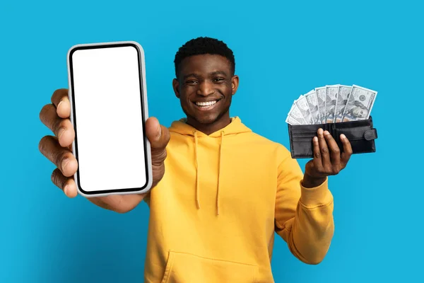 stock image Easy money, cashback. Cheerful happy handsome young african american guy in casual showing big smartphone with white blank screen and wallet full of cash dollar banknotes, blue background, mockup