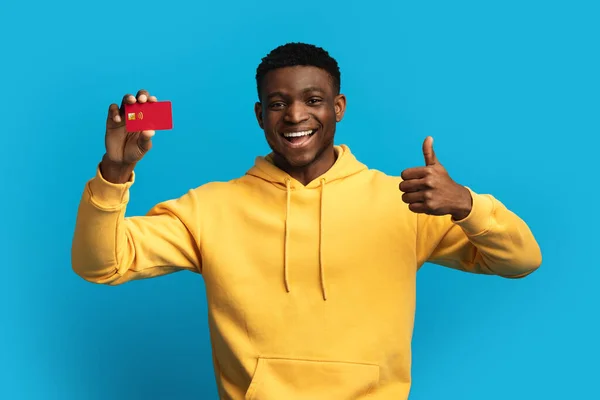 陽気なハンサムな千年黒男で黄色パーカーショー赤プラスチック銀行カードと親指アップ 非接触支払いをお勧めします 簡単に仮想銀行 青スタジオの背景 コピースペース — ストック写真