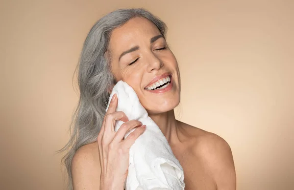 面部皮肤护理和卫生 快乐的老年妇女洗完脸后用毛巾擦干干净的脸 浴室背景是米黄色的 晨曦日常美感概念 — 图库照片