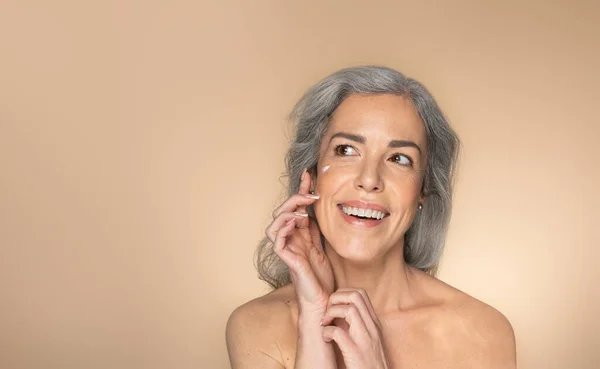 Glückliche Seniorin Die Gesichtscreme Aufträgt Ihr Aussehen Auf Beigem Studiohintergrund — Stockfoto