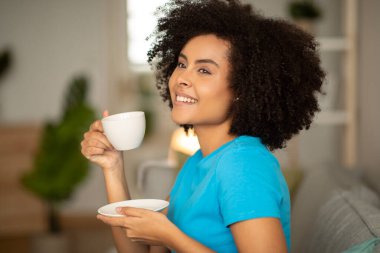 Evde günaydın. Gülümseyen milenyum kıvırcık siyah kadın elinde bir fincan çayla, rahat oturma odasındaki kanepede keyfine bakar. Akşam kahve molası, dinlenme, evde dinlenme.