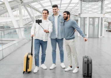 Hava Seyahat Ekibi. Bavullu üç blogcu modern havaalanının kalkış kapısında akıllı telefondan selfie çekiyor. Yolcu grubu uçuşu beklerken eğleniyor. Tam Uzunluk