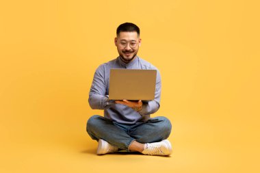 Sarı Stüdyo Arkaplanı Üzerinde Otururken Dizüstü bilgisayar kullanan Mutlu Asyalı Adam, Başarılı Millennial Erkek Serbest Yazıcı veya Blogcu Adam Bilgisayarda İnternet Taraması Yapıyor,