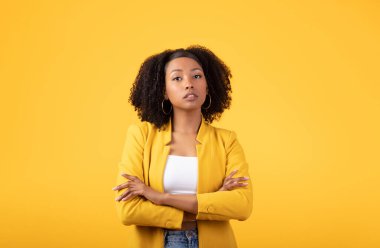 Ciddi bir siyah kıvırcık kadın portresi katlanmış kollarıyla, güzel bir Afrikalı Amerikalı kadın kameraya bakarken sarı stüdyo arka planında poz veriyor.