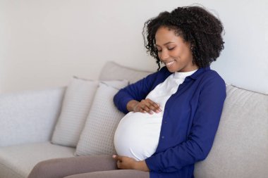 Koca göbekli, yerli giysili kıvırcık siyah bir kadının hamileliğin ve yeni hayatın tadını çıkarmasına, oturma odasındaki koltukta oturmasına sevindim. Sağlık, annelik ve evde çocuk beklentisi
