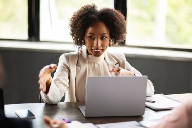 Güzel siyah kadın çalışan iş toplantısına ya da iş arkadaşlarıyla brifinge katılıyor, dizüstü bilgisayarın önünde oturuyor, konuşuyor, el kol hareketi yapıyor, yaratıcı iş fikirlerini, ofis içini paylaşıyor.