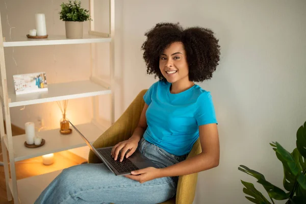 Cheerful millennial curly black lady typing on computer, watching video in living room interior. Work, freelance, business and study remotely, device and social networks at home