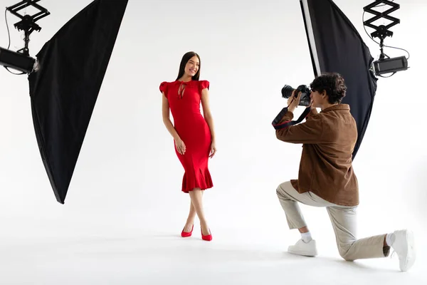 专业的年轻摄影师用现代照明设备拍摄身着红衣服的优雅女士的画像 在摄影棚的白色背景上摆出女性的照片 — 图库照片