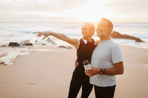 在室外海滩上 穿着运动服的成熟的高加索男人和女人戴着耳机微笑 瓶装水将手指指向空旷的空间 一起锻炼 夏天的保健和身体护理 — 图库照片