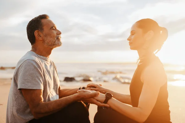 Happy calm mature caucasian man and woman in sportswear sit, practice yoga, meditate in morning on beach, outdoor. Couple enjoy workout together, peace, health care, body care in summer