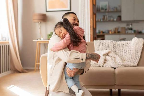 Joy Of Fatherhood. Loving Asian Dad Embracing His Cute Little Baby Daughter, Expressing Care And Love Posing In Modern Living Room At Home. Family Happiness, Cute Moments Concept