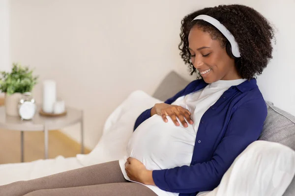 ワイヤレスヘッドフォンで大きな腹を持つ陽気な巻きミレニアルブラック女性は妊娠を楽しみ 寝室のインテリアで音楽を聴く 出産の準備 母親と子供の期待 — ストック写真