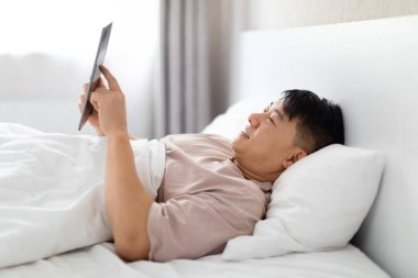 Akıllı ev, modern teknoloji konsepti. İyi dinlenmiş yakışıklı orta yaşlı Asyalı adam yatakta dijital tablet kullanıyor, ekranlara dokunuyor, perdeleri açıyor, klimayı ayarlıyor, yan görüş, boş alan.