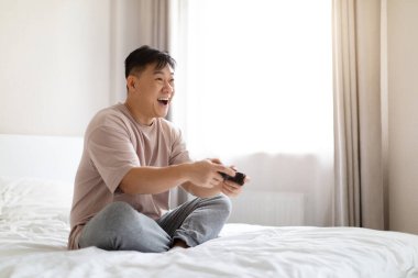 Evde dinlenmek, ev eğlencesi kavramı. Heyecanlı orta yaşlı Çinli adam evde yatağında oturuyor, elinde joystick tutuyor ve bağırıyor, video oyunları oynuyor, oyunu kazanıyor, uzayı kopyalıyor.