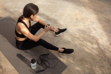 Egzersiz sonrası dinlenen yorgun, beyaz tenli bayan, atlama halatı ve su şişesiyle mindere oturmuş, nabzını kontrol ediyor. Spor, eğitim uygulaması, fitness, kilo kaybı, sağlık hizmetleri