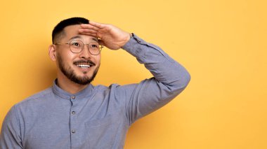 Gülümseyen Asyalı adam, elini alnına yaklaştırıyor ve ilgi ile fotokopi uzayına bakıyor, neşeli, yakışıklı erkek, sarı arka planda dururken teklifin tadını çıkarıyor.