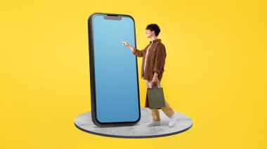 İnternetten alışveriş uygulaması. Neşeli Alıcı Adam, sarı stüdyo arka planında alışveriş çantasının yanında duran boş ekranlı, büyük cep telefonu kullanıyor. Kolaj, Panorama