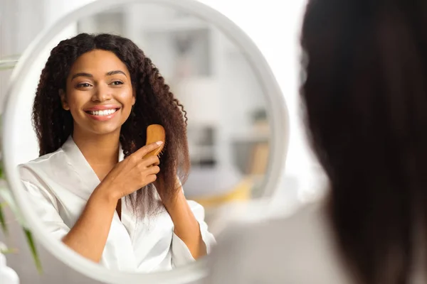 Haircare Routine. Attractive Black Woman Combing Her Beautiful Curly Hair With Bamboo Brush At Home, Happy Young African American Lady Looking To Mirror And Smiling, Selective Focus On Reflection