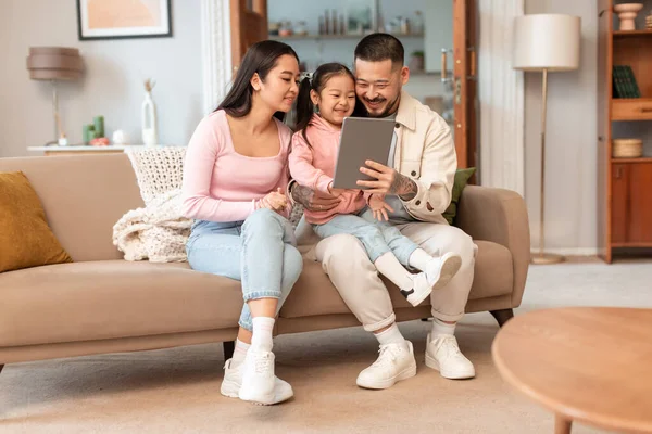 家族オンラインレジャー 韓国人の両親と小さな娘は 現代のリビングルームの屋内で一緒に座ってコンピュータ上で漫画をオンラインで見てタブレットを使用して インターネットと楽しいコンセプト — ストック写真