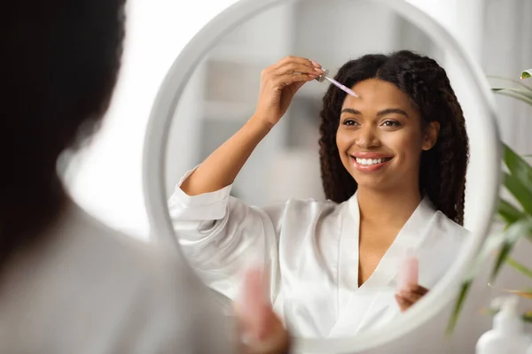 スキンケアコンセプト 美しい黒の女性がミラーの近くに顔の血清を適用します 自宅で毎日のスキンケアルーチンを作る幸せなアフリカ系アメリカ人女性 新しい化粧品をテスト 反射に選択的な焦点 — ストック写真