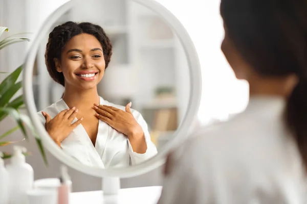 鏡を見ながらデコルテゾーンに触れる黒人笑顔の女性 白い絹のローブを着て首にしわをチェックするアフリカ系アメリカ人女性 アンチエイジング治療の結果を楽しむ — ストック写真