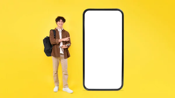 在大型Cellphone附近的快乐学生家伙展示空白屏幕和应用程序广告的自由空间 在黄色工作室背景下拥抱现代技术和背包 — 图库照片