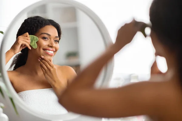 若いアフリカ系アメリカ人女性のマッサージ顔とともにGua Shaクォーツ石スクレーパースキンケア 鏡を見て黒の女性の笑顔 自宅で美容トリートメントを作り 健康な輝く肌を楽しむ — ストック写真