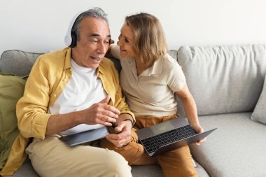 Aged senior man listening to music in wireless headphones, woman raising the earpiece and talking in husbands ear, sitting on sofa at home, free space clipart
