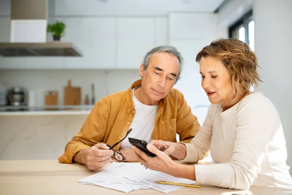 Senior couple sitting in kitchen with papers and bills and using calculator, husband and wife counting monthly expenses or planning family budget at home