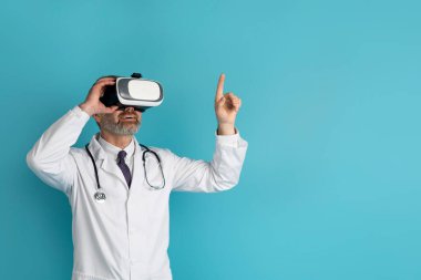 Doktor orta yaşlı, sanal gerçeklik gözlüğü takan, mavi stüdyo arka planında izole edilmiş, fotokopi alanına dokunan adam. Sağlık hizmetleri kavramında geleceğin tıbbı ve modern teknoloji aletleri