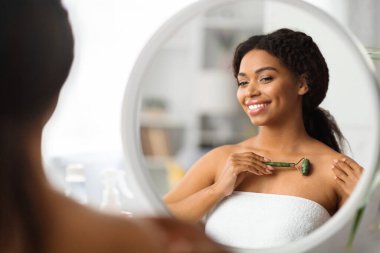 Güzellik Tedavileri. Gülümseyen Siyah Kadın Yeşiltaş Patenini Evde Kullanıyor, Mutlu Afrikalı Amerikalı Kadın Banyoda Aynaya Bakarken Vücut Kaldırıyor, Seçici Odaklanma